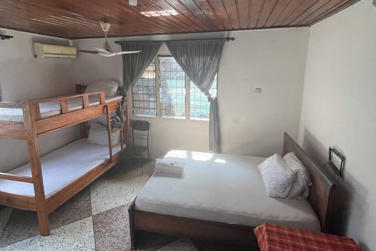 Une chambre avec un lit superposé, un lit simple, une petite table recouverte d’une nappe à carreaux rouges et une fenêtre avec des rideaux.