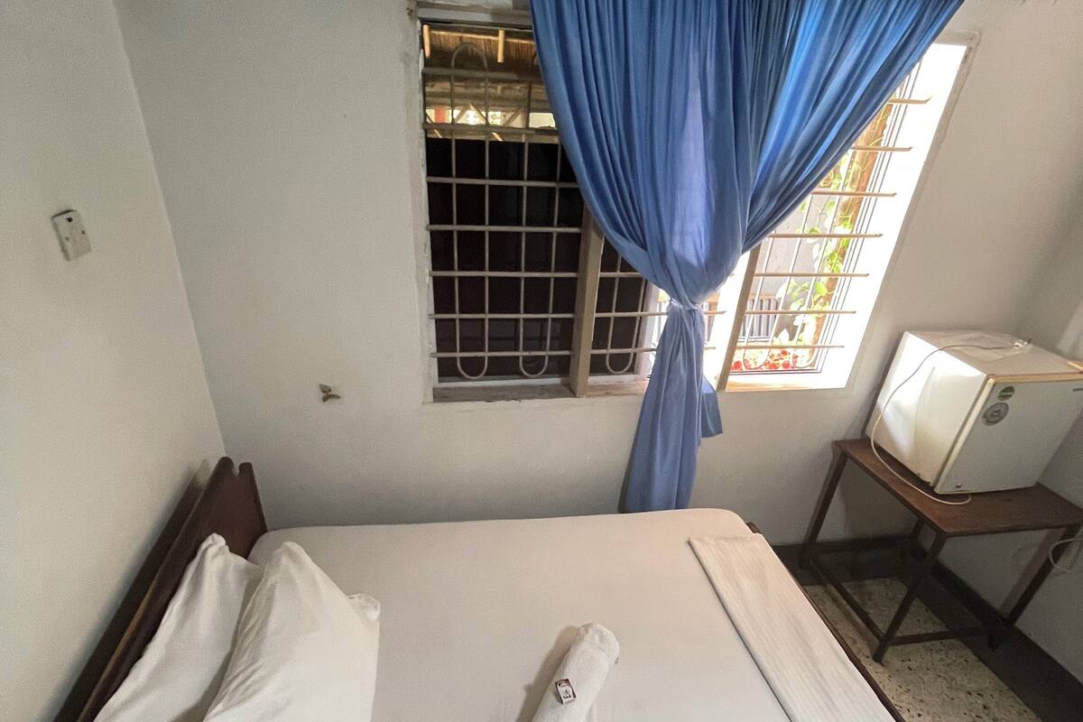 Un lit simple avec du linge de lit blanc, une serviette enroulée et un oreiller, dans une pièce dotée d’une fenêtre recouverte de rideaux bleus.