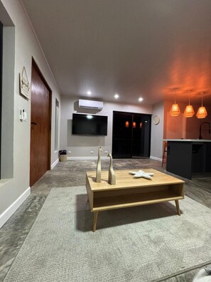 Ein modernes Wohnzimmer mit Holztisch, Flachbildfernseher an der Wand und Küchenzeile mit Pendelleuchten.