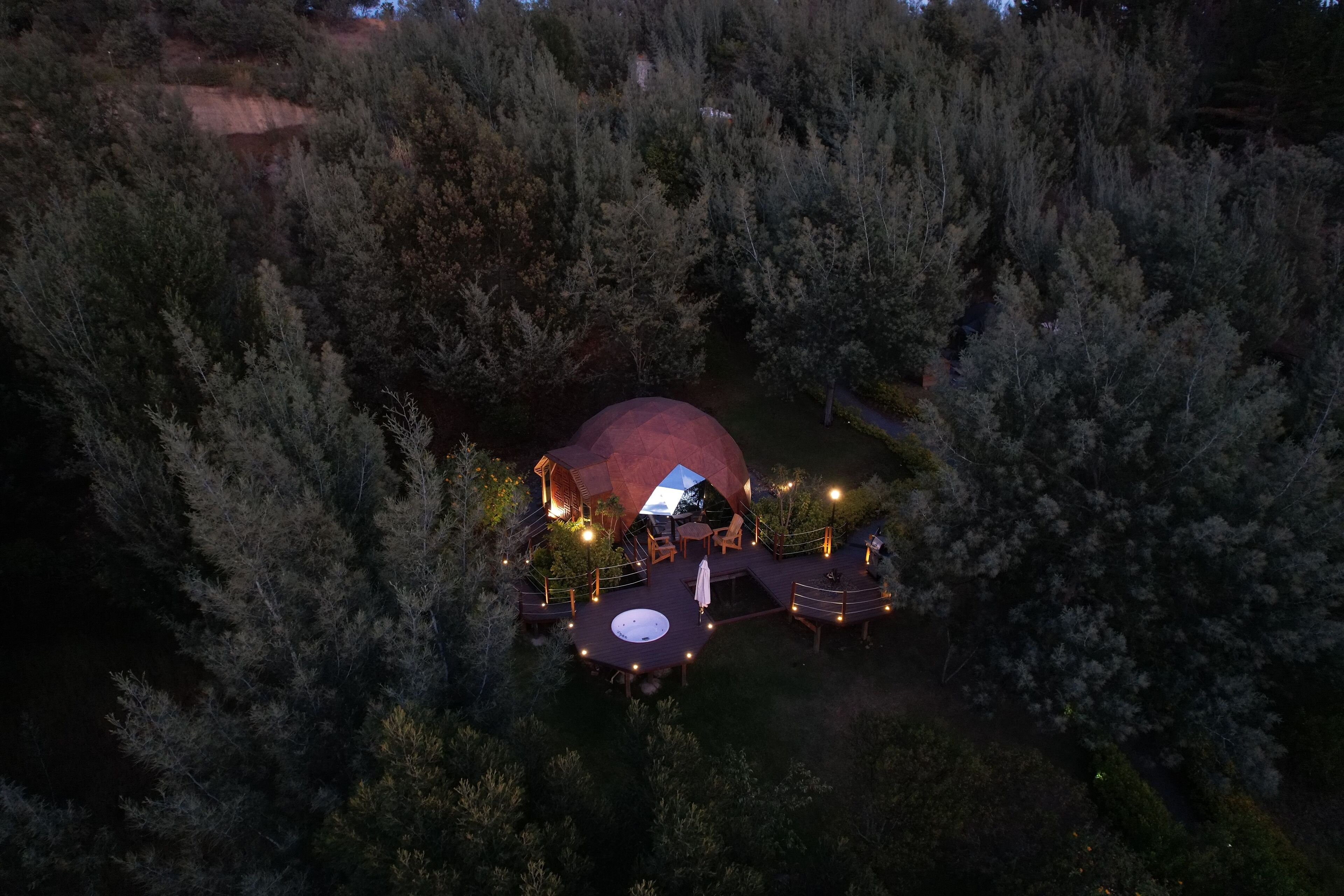 Vista aérea de una estructura en forma de cúpula rodeada de un bosque denso, con una terraza y un área de estar al aire libre.
