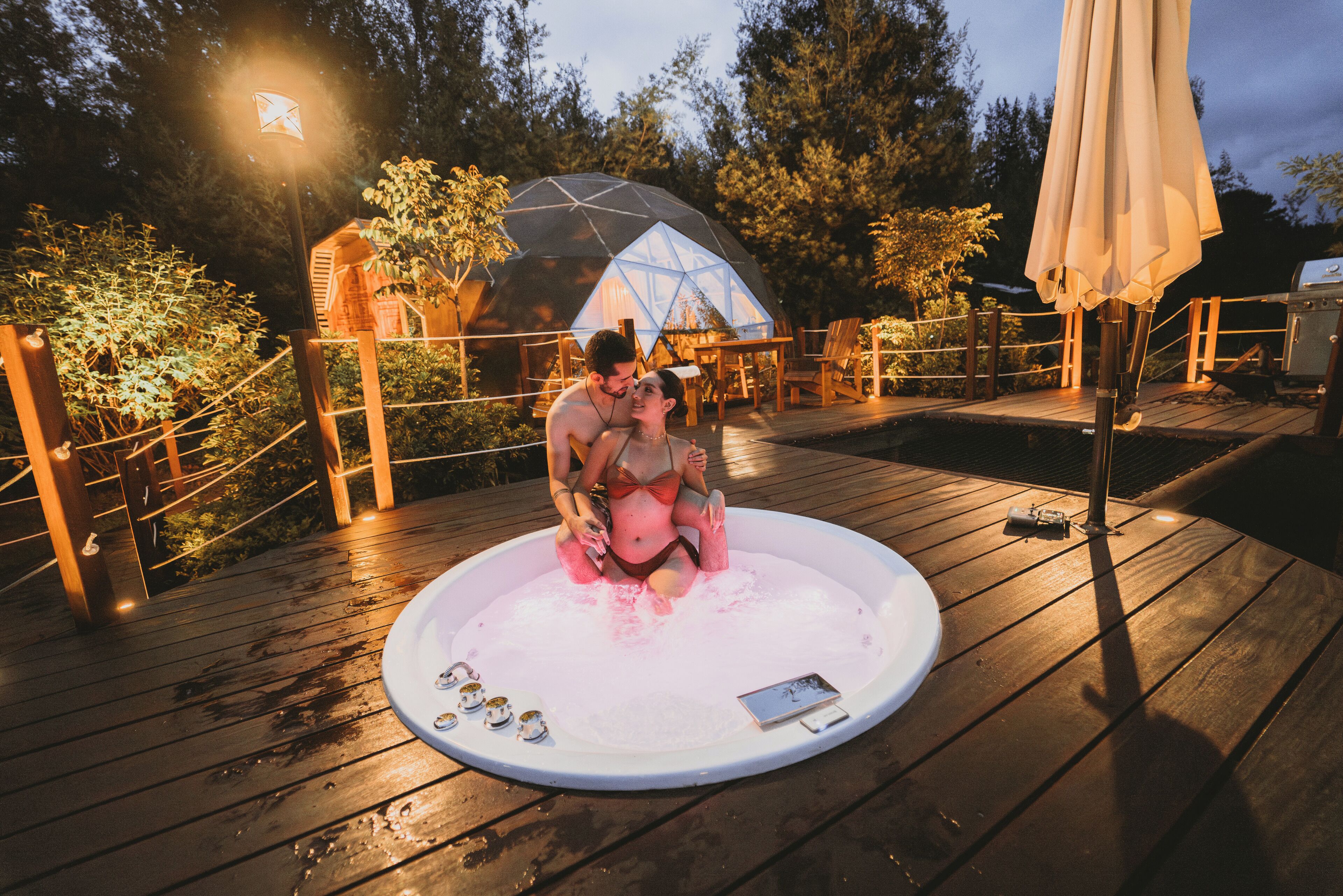 A couple in a hot tub on a wooden deck with a geodesic dome structure in the background.