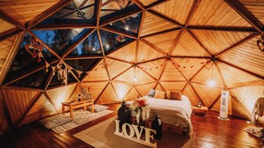 A cozy, triangular wooden tent with a bed, a small table, and a 'LOVE' sign in the center. - Ráquira Glamping (Ráquira)