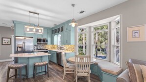 A kitchen with teal cabinets, a stainless steel refrigerator, a microwave, and a stove. There is a dining area with a round table and chairs, and a window with blinds. - Redfish Cottage - Old Florida Cottages, Community Pool, Easy Walk to the Beach! (Old Florida Cottages)
