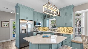 A kitchen with teal cabinets, a stainless steel refrigerator, a microwave, and a large island with a bowl and a plate. - Redfish Cottage - Old Florida Cottages, Community Pool, Easy Walk to the Beach! (Old Florida Cottages)