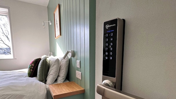 A bedroom with a bed, a bedside table, a wall-mounted control panel, and a window.