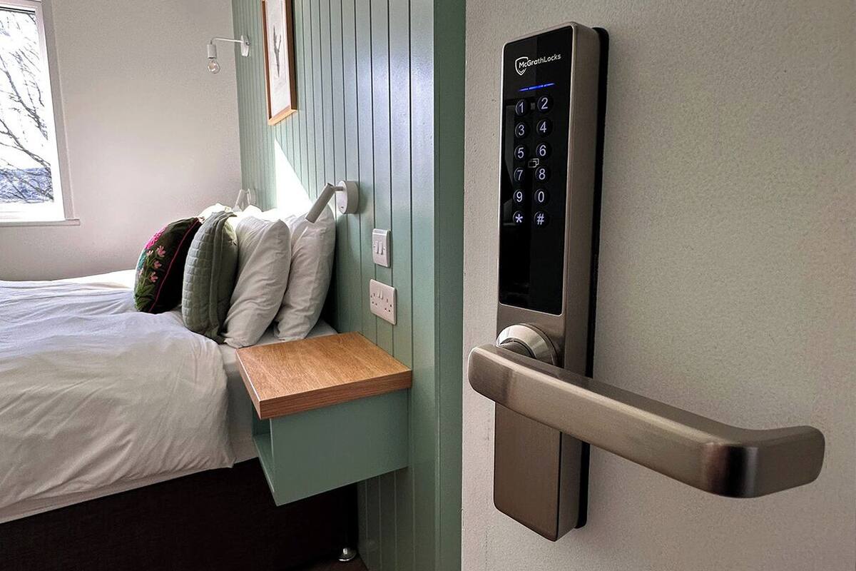 A bedroom with a bed, a bedside table, a wall-mounted control panel, and a window.