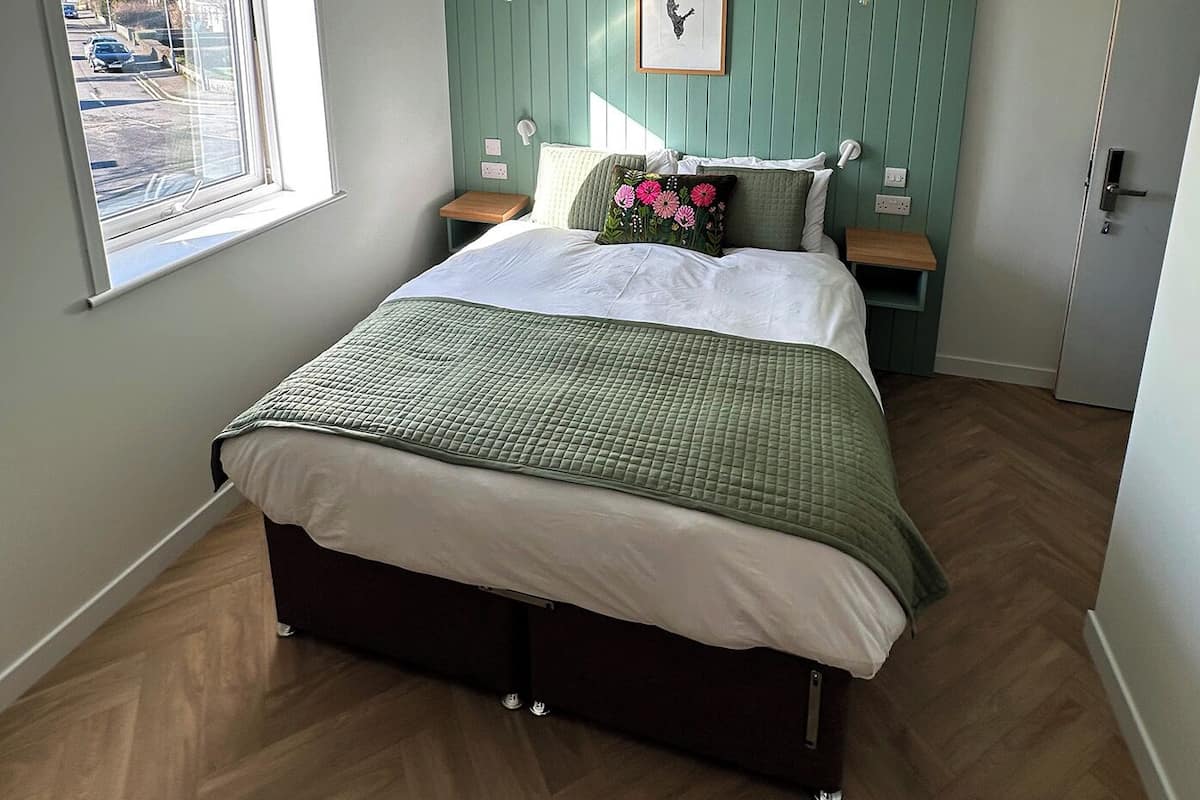A bedroom with a bed, a window, a bedside table, a nightstand, a framed picture, and a door.