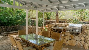 An outdoor patio with a glass-top table surrounded by wicker chairs, a stone wall, and a built-in grill. - Private 2 Bedroom Home | Pool | Near Beach (Coco)