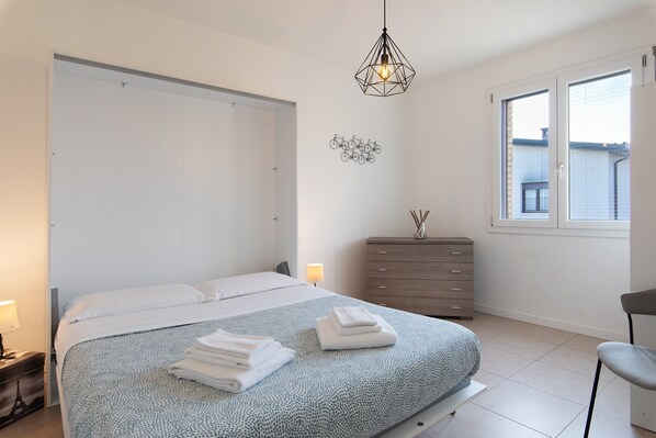 Une chambre à coucher comprenant un lit, une commode, une chaise, une fenêtre et un luminaire fixé au mur.