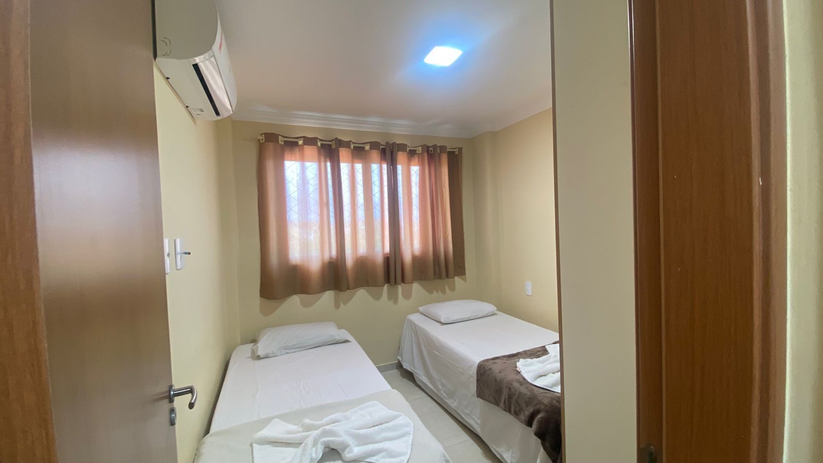 Una habitación pequeña y limpia con dos camas, una ventana con cortinas, un equipo de aire acondicionado y una puerta.