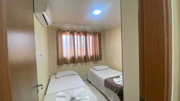 Una habitación pequeña y limpia con dos camas, una ventana con cortinas, un equipo de aire acondicionado y una puerta.