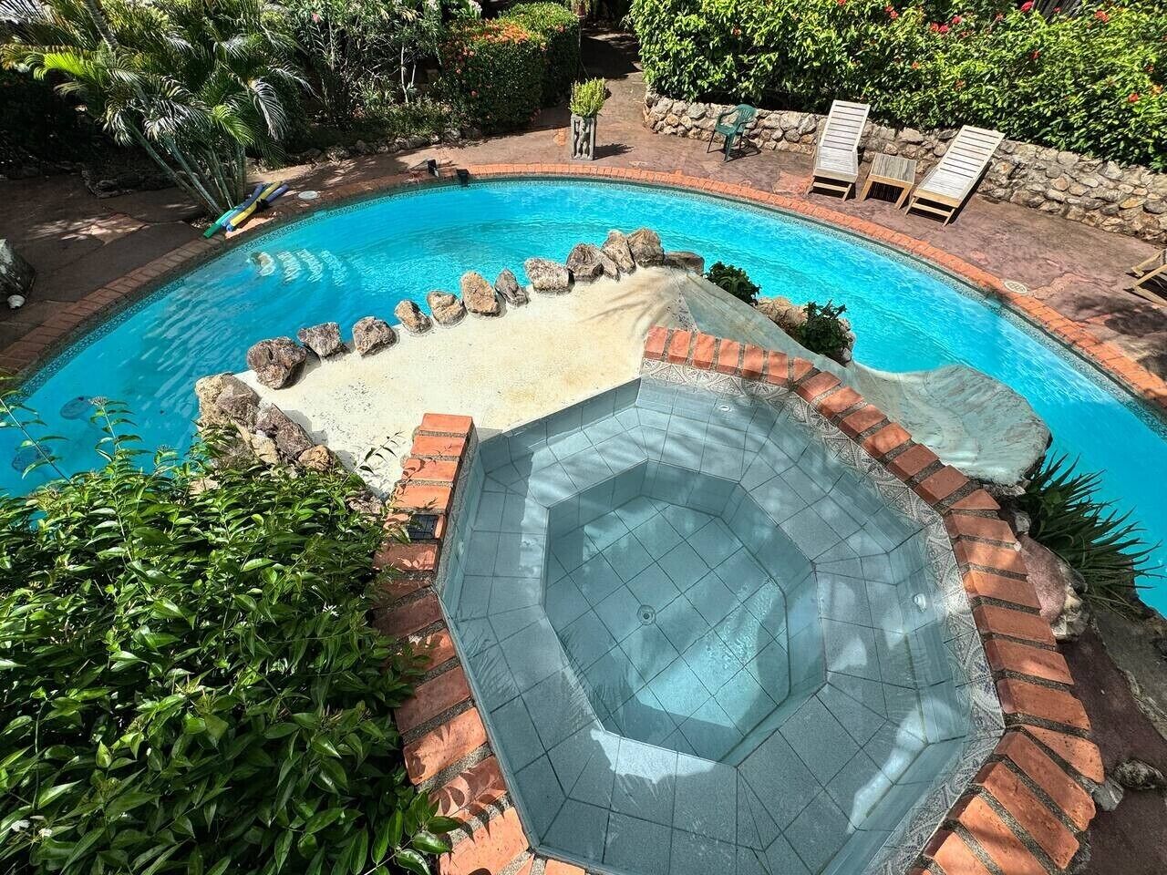 En swimmingpool med ett bubbelbad, omgiven av en terrass av tegelsten och grönska.