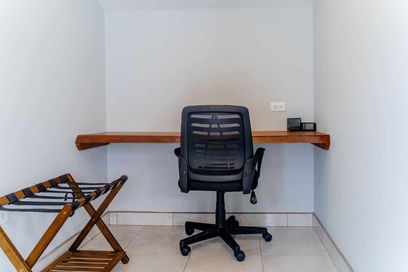 A corner office with a black office chair, a wooden desk, a small shelf with a black object, and a wooden chair with a black seat.
