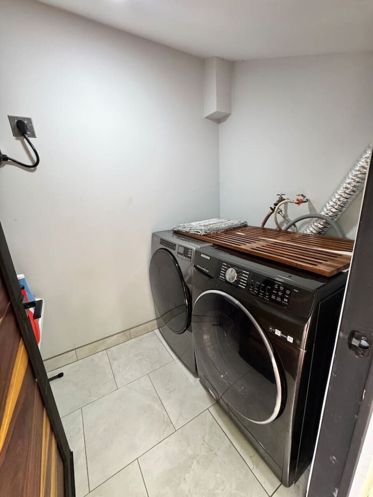 Une petite buanderie équipée d’un lave-linge et d’un sèche-linge à chargement frontal, d’un étendoir en bois et d’une bouche d’aération fixée au mur.