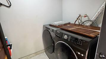 Une petite buanderie équipée d’un lave-linge et d’un sèche-linge à chargement frontal, d’un étendoir en bois et d’une bouche d’aération fixée au mur.