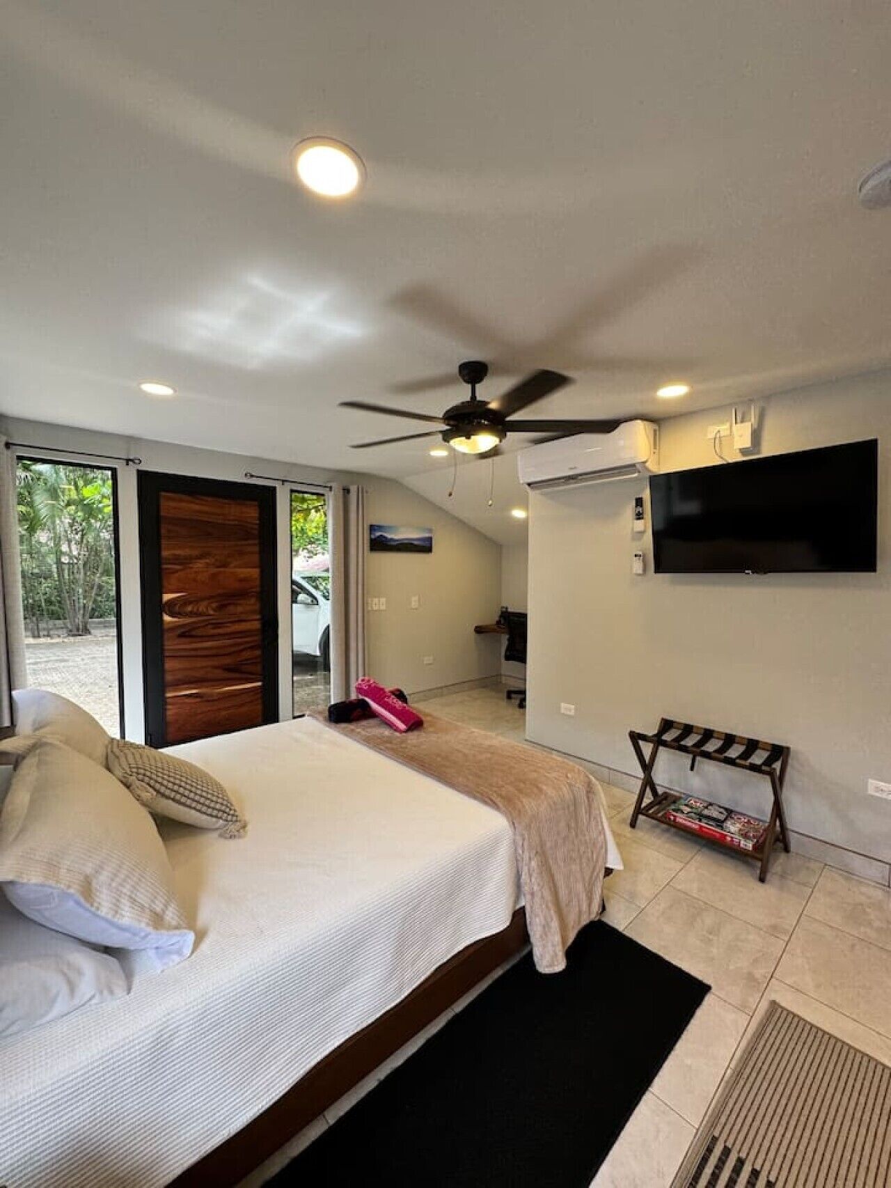 A bedroom with a bed, a ceiling fan, an air conditioning unit, a TV mounted on the wall, and a view of the outside.
