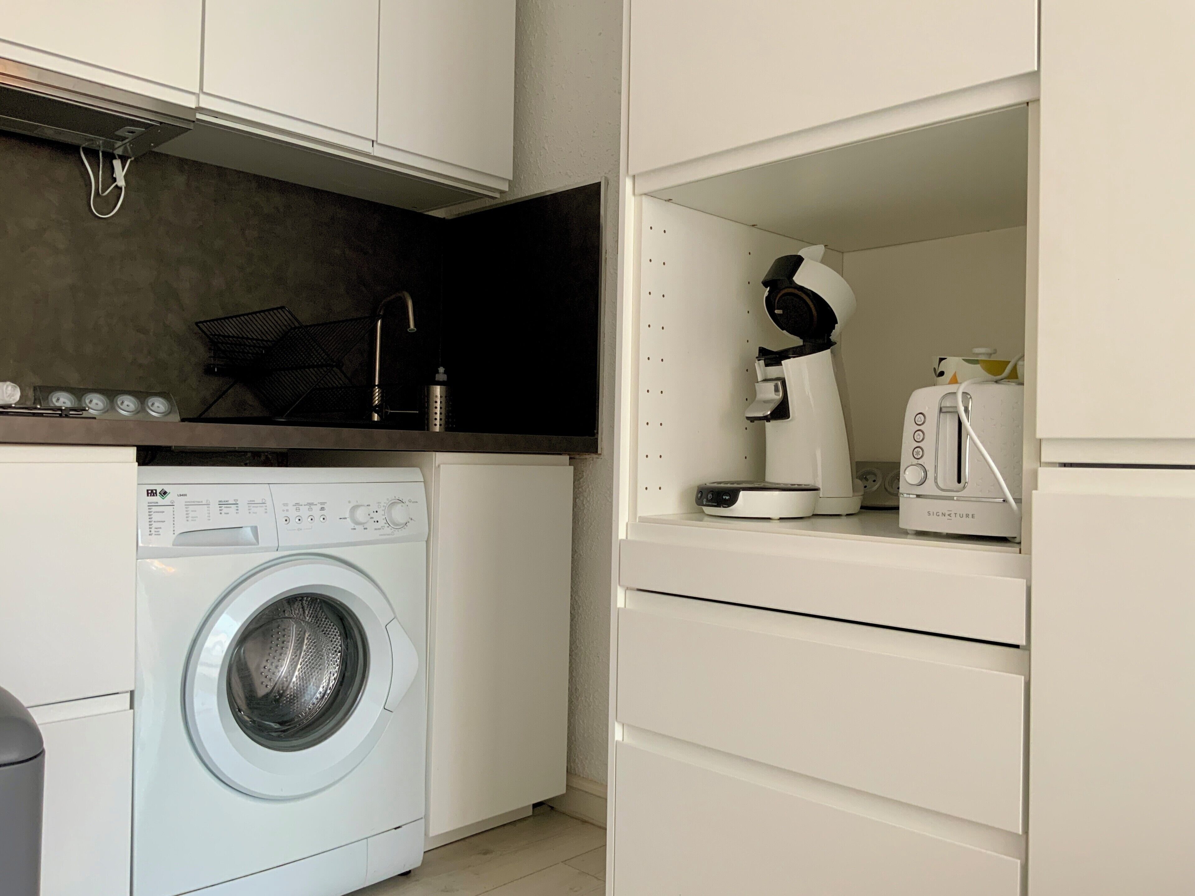 A compact kitchen with a front-loading washing machine, a coffee maker, and a toaster.