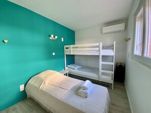 Ein Zimmer mit türkisfarbenen Wänden, einem Etagenbett, einem Einzelbett mit zusammengerolltem Handtuch, einem kleinen Tisch und einer Klimaanlage.