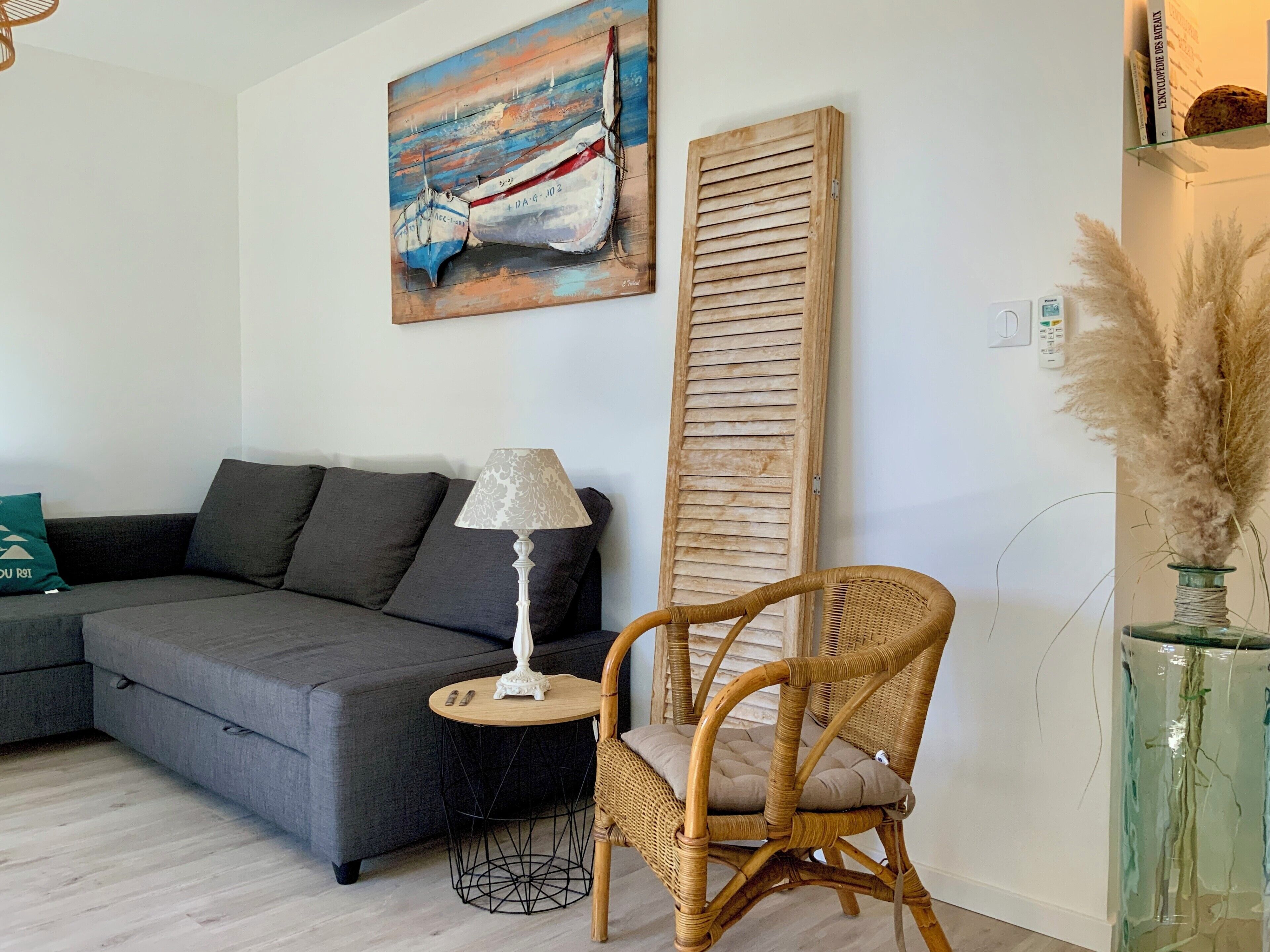 Ein Wohnzimmer mit grauem Sofa, einem Korbstuhl, einem kleinen runden Tisch mit Lampe, einem Gemälde von Booten, einem Holzklappladen und einer Vase mit Trockenpflanzen.
