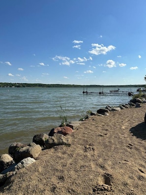 Blick auf einen See mit sandigem Ufer, Felsen und Booten auf dem Wasser.