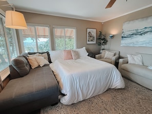 A bedroom with a large bed, a sofa, and a chair. There are windows with striped curtains, a ceiling fan, and a painting on the wall.