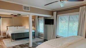 Une chambre à coucher avec un lit, un ventilateur de plafond, une télévision et une grande fenêtre équipée de stores.