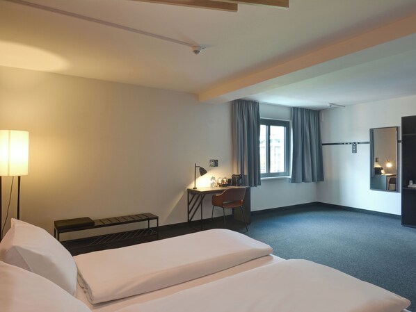 Habitación de hotel con cama, escritorio, silla, lámpara y ventana con cortinas.