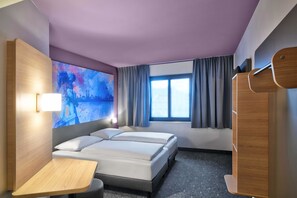 Habitación de hotel con dos camas, un escritorio, un ventanal grande con cortinas y un televisor instalado en la pared.
