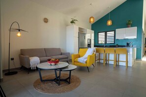Ein modernes Wohnzimmer mit einer Couch, einem gelben Sessel, einem runden Tisch mit Obstkorb und einer Küchenzeile mit Bar.