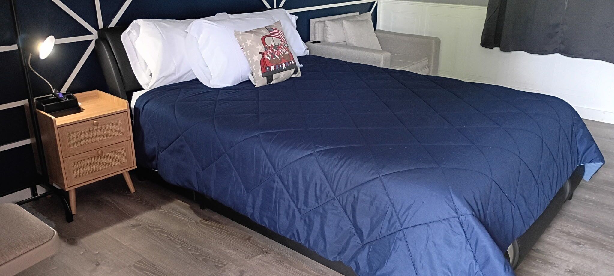 Una cama con colcha azul oscuro, una mesita de noche con lámpara y una pared con patrón geométrico.