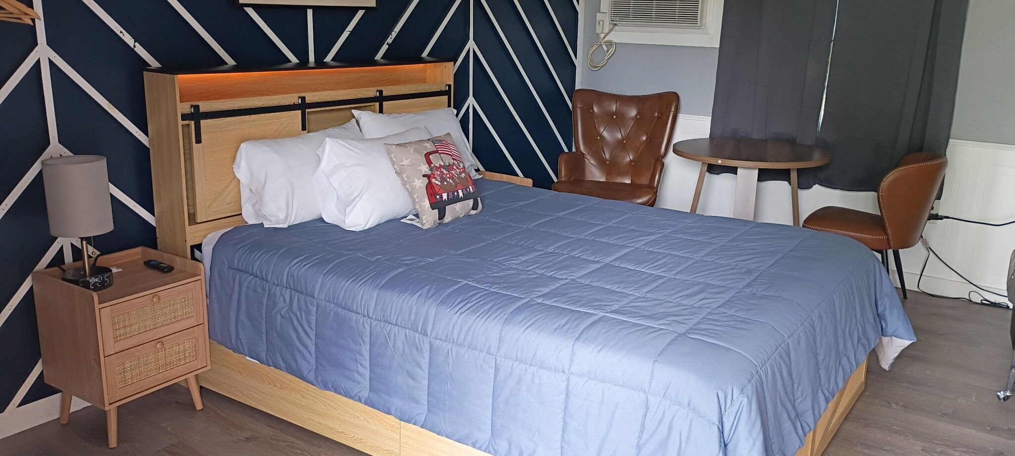 Un lit bien fait, recouvert d’une couvre-lit bleue, une table de chevet en bois avec une lampe, un fauteuil en cuir marron et une petite table ronde avec une chaise.