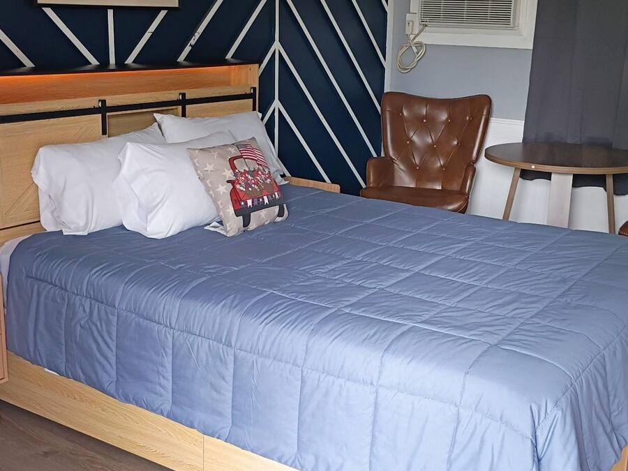 Un lit bien fait, recouvert d’une couvre-lit bleue, une table de chevet en bois avec une lampe, un fauteuil en cuir marron et une petite table ronde avec une chaise.