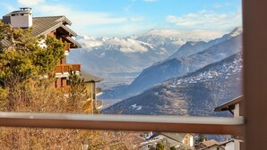Vista de montanha a partir de uma varanda, com uma casa de madeira e picos nevados ao fundo.
