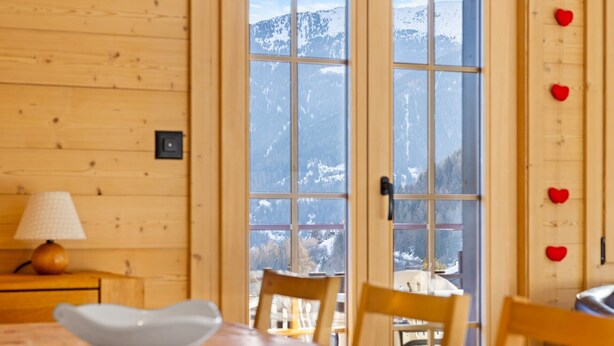 Interior em madeira com uma mesa, cadeiras, uma lâmpada e vista para montanhas cobertas de neve através de janelas amplas.