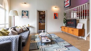 Ein Wohnzimmer mit Sofa, Couchtisch und Fernseher. Es gibt ein Bücherregal, eine Lampe und ein Schild mit der Aufschrift 'Fish and Chips'.