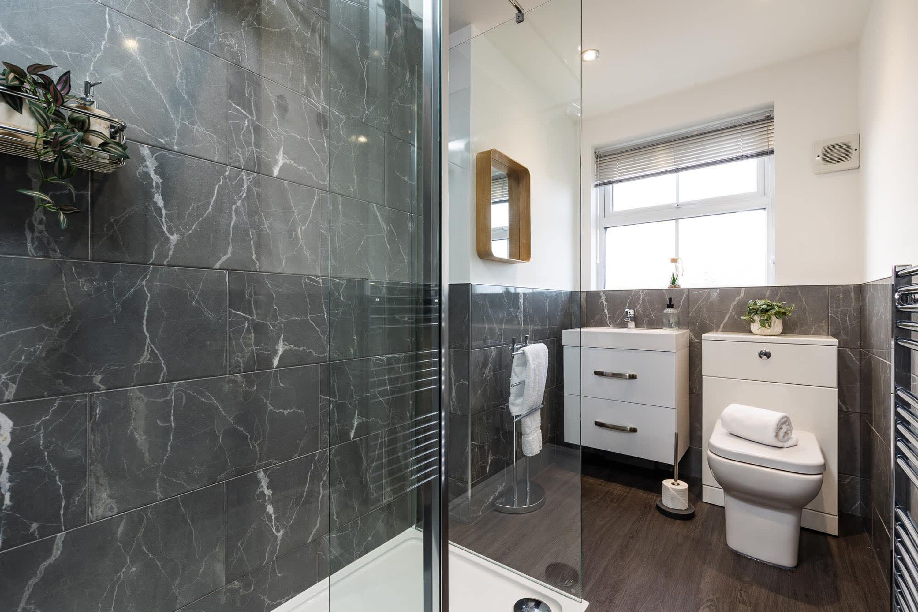 A modern bathroom with dark marble tiles, a glass shower enclosure, a white toilet, and a small bedside table with a plant.