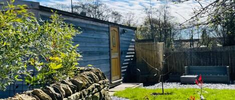 A rustic cabin with a stone wall, a wooden door, and a gravel path leading to a grassy area with a wooden bench.