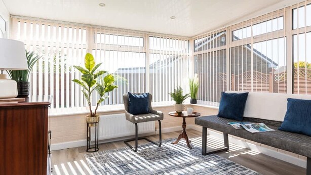 Een zonnige kamer met een bank, fauteuil en een klein tafeltje, met grote ramen en potplanten.