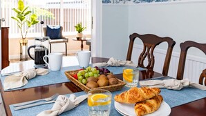 Een ontbijttafel met croissants, fruit en sapglazen, geplaatst op een blauwe mat in een goed verlichte kamer met houten stoelen en uitzicht op het buiten.