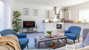 Uma sala de estar moderna com um sofá, duas poltronas, uma mesa de centro e uma TV exibindo 'NETFLIX'.