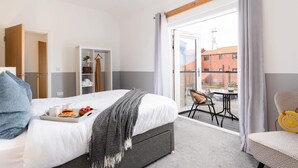 Quarto de hotel com uma cama, uma mesinha com uma cadeira, um guarda-roupa e uma varanda com vista para edifícios.