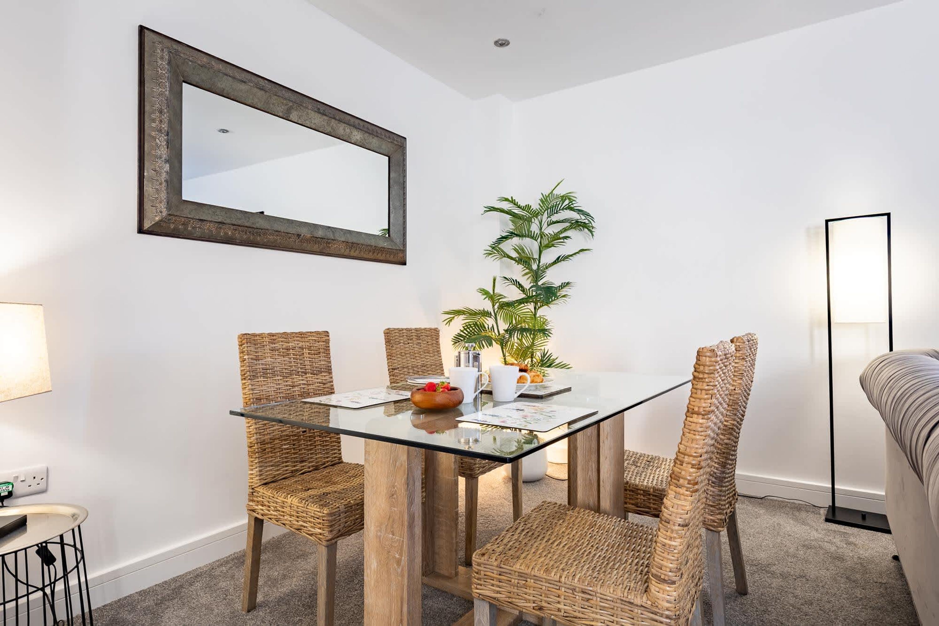 A dining area with a glass table, wicker chairs, a potted plant, and a mirror on the wall.