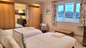 Ein Hotelzimmer mit zwei Betten, einem Holzschrank, einem Stuhl mit Kissen, einer Holzbank und einem Fenster mit Vorhängen.
