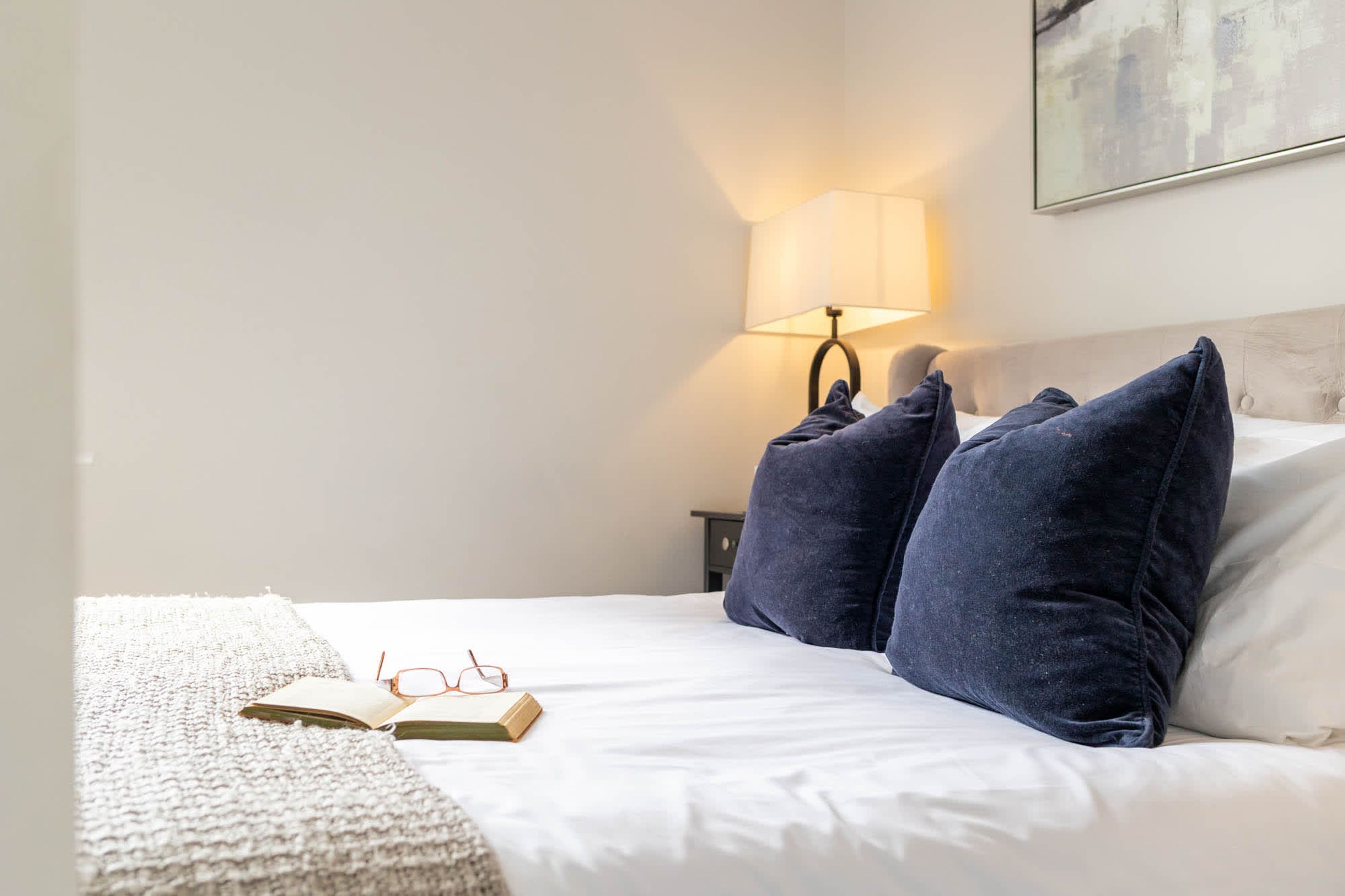 A neatly made bed with a white comforter and two dark blue pillows, a pair of glasses, and an open book on a textured blanket.