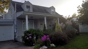 A two-story house with a covered porch, a garage, and a well-maintained front yard with flowering plants. - Cozy 1-bedroom on the 2nd floor in Valley Stream, with a beautiful outdoor deck. (Valley Stream)