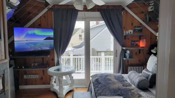 A cozy room with a bed, a television, a small table, and a view of the outside. - Cozy 1-bedroom on the 2nd floor in Valley Stream, with a beautiful outdoor deck. (Valley Stream)