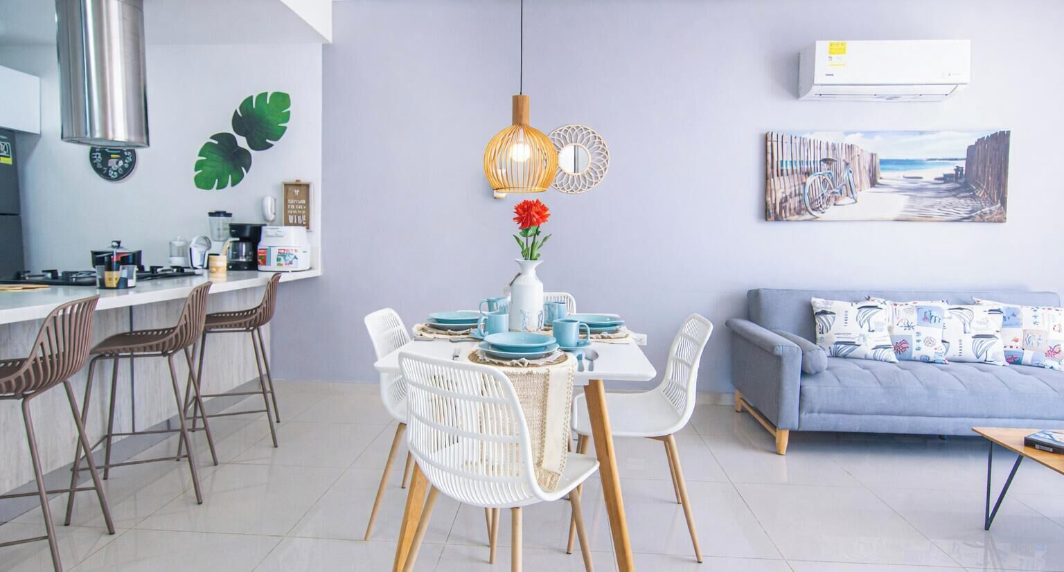 A modern kitchen with a dining area, featuring a table set with blue plates and white chairs, a vase with red flowers, and a painting of a beach scene on the wall.