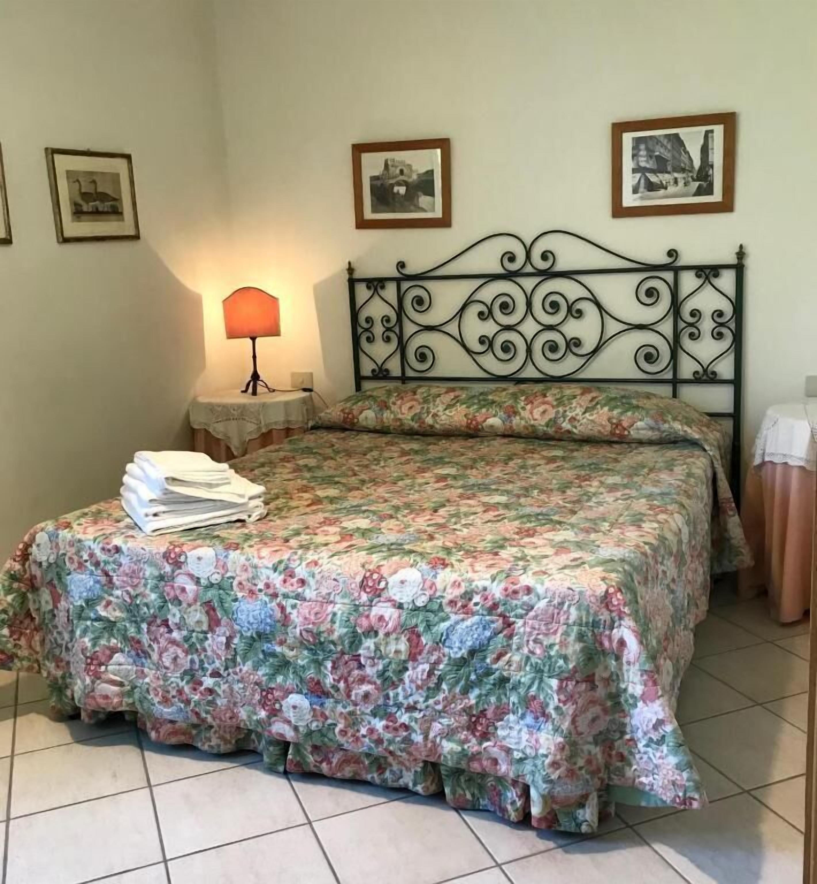 A bedroom with a floral bedspread, a metal headboard, a bedside lamp, and two framed pictures on the wall.