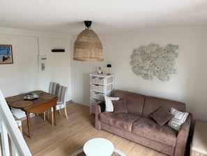 Een gezellige woonkamer met een bruine bank, een eettafel met stoelen, een rieten hanglamp en een visdecoratie aan de muur.