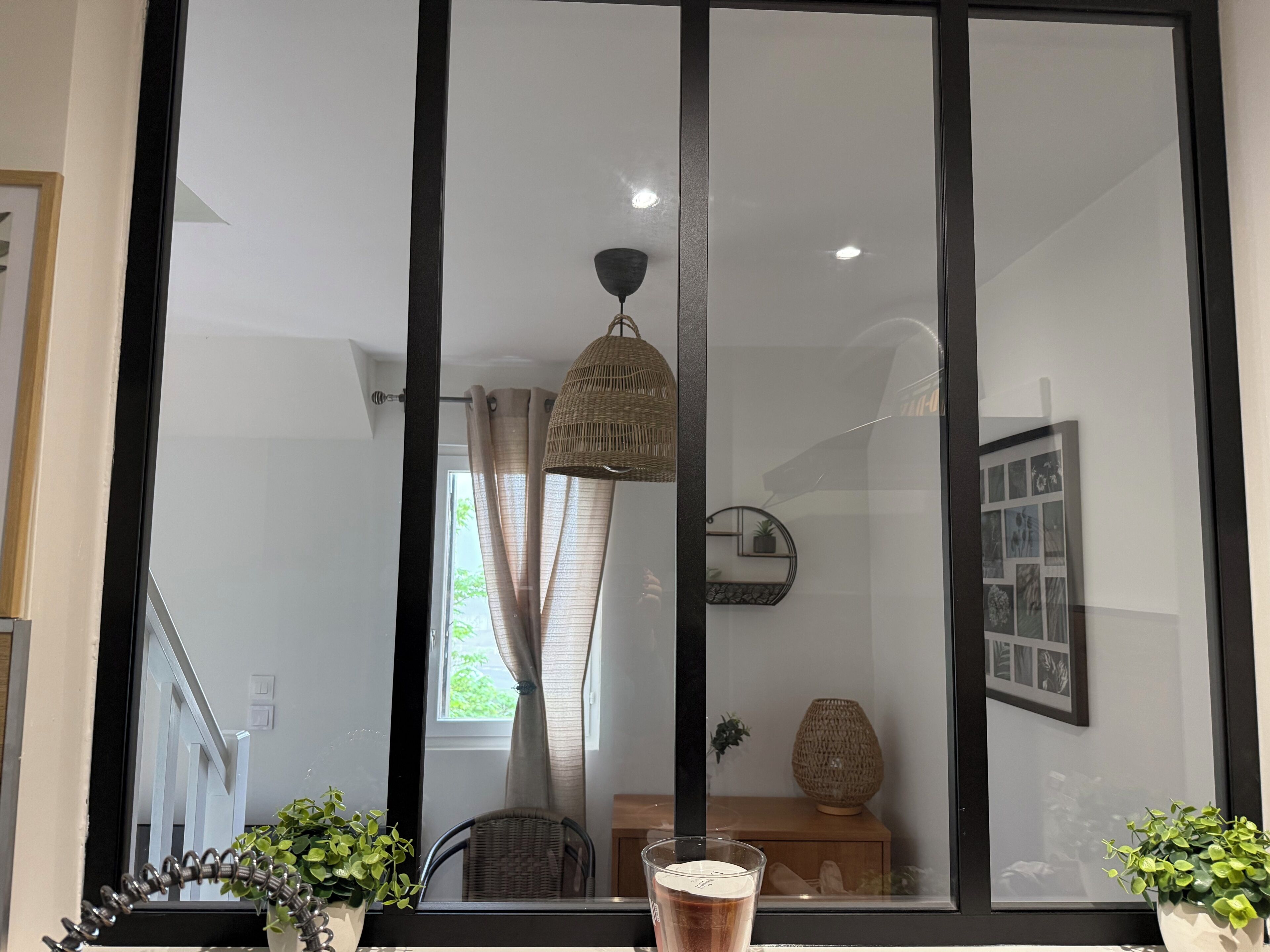 Quarto com uma janela grande, uma cadeira de vime, uma mesa pequena e um vaso com planta.